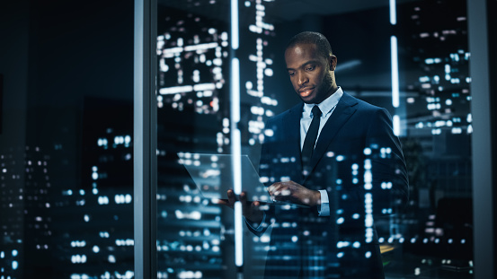 A Black Businessman working on his laptop and ooking out of window of a city late at night.